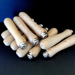 Puede incluir: Un conjunto de mangos de madera con férulas metálicas. Los mangos son de color marrón claro, cilíndricos y cónicos, con un acabado liso. Las férulas metálicas son de color plateado y rodean la base de cada mango. Los artículos están dispuestos sobre una superficie negra.