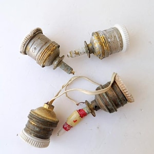 Puede incluir: Cuatro casquillos de bombilla de metal vintage con vástagos roscados y anillos de plástico blanco. Dos casquillos están conectados por un cable eléctrico blanco, mientras que los otros dos están separados. Las superficies metálicas muestran desgaste.
