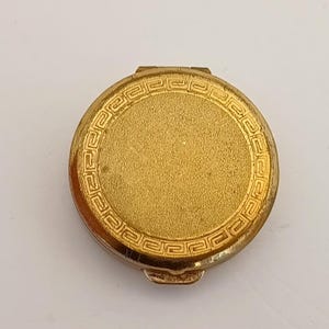 May include: A circular, gold-coloured compact with a textured surface and a decorative border. The compact is closed and has a small clasp. The design is simple and elegant, suitable for carrying makeup or other small items.