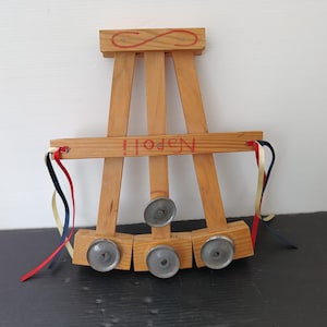 May include: Wooden toy with a light brown finish, featuring an easel design with three small wheels. The word "Napoli" is painted in red. Red, blue, and yellow ribbons are attached to the sides. An infinity symbol is painted on the top.
