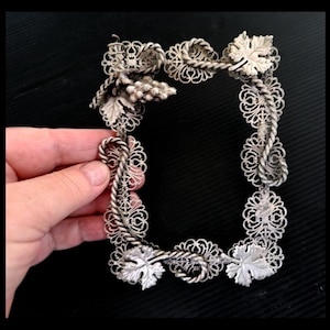 May include: A silver-coloured, rectangular picture frame with detailed filigree work. The frame features grape clusters and leaves, forming a vine-like pattern. The frame is held by a hand against a dark backdrop.