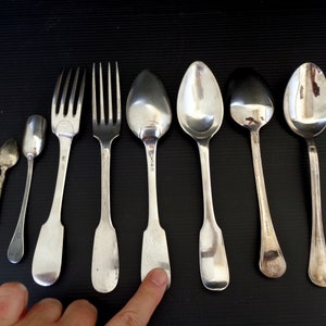 May include: A set of seven silver-plated utensils, including two forks, four spoons, and a small spoon. The utensils are arranged in a row on a black surface.