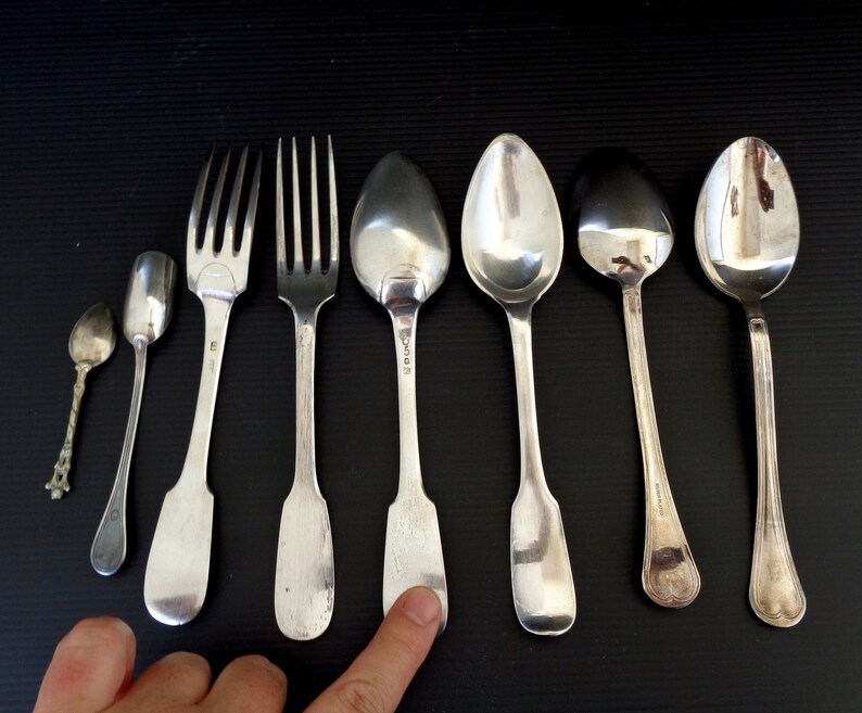 May include: A set of seven silver-plated utensils, including two forks, four spoons, and a small spoon. The utensils are arranged in a row on a black surface.