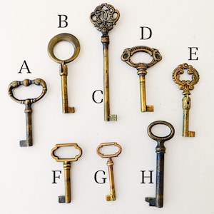 May include: A collection of eight antique-style metal keys labeled A through H. Each key features a distinct bow shape, ranging from simple circles to ornate scrollwork, with a weathered brass finish and a standard bit at the bottom of the shaft.