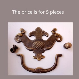 Pode incluir: Um conjunto de cinco puxadores de armário de latão de estilo antigo. Os puxadores apresentam um design decorativo e ornamentado com um centro em forma de abacaxi. O conjunto inclui dois puxadores com uma alça curva e duas maçanetas redondas menores.