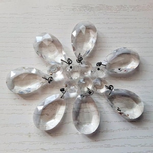 Op de afbeelding: Heldere kristallen kroonluchterdruppels, gerangschikt in een cirkelvormig patroon. Elke druppel is druppelvormig met meerdere facetten, verbonden door kleine metalen lussen. De kristallen reflecteren licht en creëren een sprankelend effect.