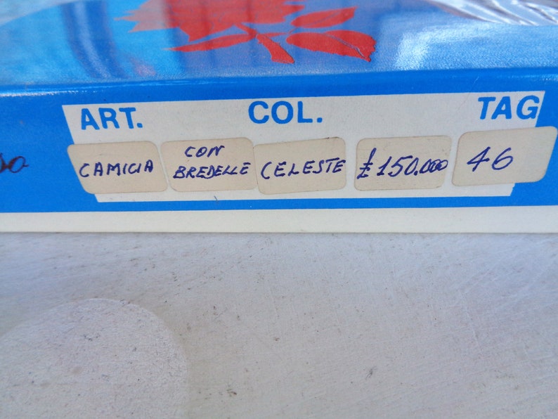 May include: A blue box with white labels that say "ART. CAMICIA CON BREDELLA CELESTE L 150.000 46"