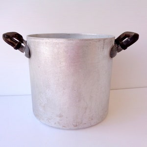Puede incluir: Una olla grande de aluminio plateado con dos asas negras. La olla tiene una superficie ligeramente desgastada.