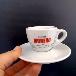 Könnte beinhalten: Weiße Espressotasse und Untertasse aus Keramik mit dem Schriftzug "Caffe Moreno" in roter Schrift. Die Tasse zeigt eine Schwarzweißillustration einer Stadtlandschaft.