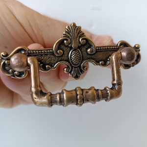 Op de afbeelding: Een sierlijke bronzen ladeknop met een decoratief middenontwerp en gebogen handvat. De hardware heeft een gedetailleerd bloemen- en scrollpatroon, perfect voor het restaureren van antieke meubels.