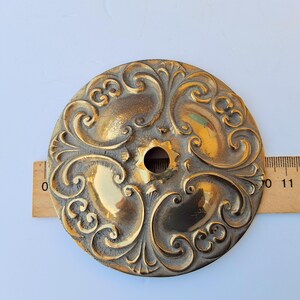 May include: A circular brass medallion featuring an ornate, raised scrollwork pattern and a central hole. A wooden ruler is positioned behind the object, indicating a diameter of approximately 4 inches across the metallic surface.