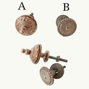 Peut inclure: Deux types de boutons de placard en métal étiquetés A et B. Le bouton A a une finition cuivrée avec un sommet pointu et étagé et des motifs en relief. Le bouton B présente une finition bronze patiné avec une face plate et des stries.