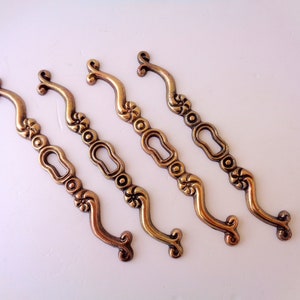 May include: Four antique brass-toned drawer pulls. Each pull features a curved handle, decorative floral accents, and a keyhole-shaped design in the center. The pulls are arranged diagonally against a white background, showcasing their vintage aesthetic.