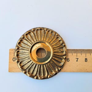 May include: A circular brass medallion with a central hole and a raised petal-like pattern radiating from the center. A wooden ruler is positioned behind the object, showing a diameter of approximately 2.5 inches across the metal piece.