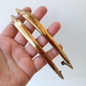 Peut inclure: Deux poignées de tiroir en laiton incurvées et allongées avec une finition métallique polie. Chaque poignée présente un design effilé et pointu aux deux extrémités, affichant une surface lisse et réfléchissante avec des variations subtiles.