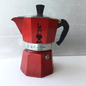 May include: A red aluminium stovetop espresso maker with a black plastic handle and lid knob. The body features the Bialetti logo of a man with a moustache and the text Moka Express Made in Italy printed on the silver-coloured middle section.