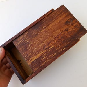 Puede incluir: Una caja rectangular de madera con tapa corredera, teñida en tonos marrones. La caja está abierta, revelando el interior. La veta de la madera es visible y la caja está sujeta por una mano.