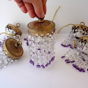 Op de afbeelding: Vintage messing en kristallen kroonluchteronderdelen. De armaturen hebben heldere en paarse kristallen kralen. De messing componenten hebben sierlijke details. De kroonluchteronderdelen liggen op een wit oppervlak.