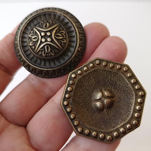 Könnte beinhalten: Zwei antike Schrankknöpfe aus Bronze. Einer ist rund mit einem floralen Design, der andere hat eine achteckige Form mit einer strukturierten Oberfläche und kleinen dekorativen Punkten am Rand. Beide haben ein Vintage-Aussehen.