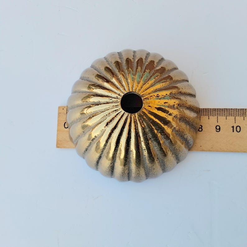 May include: A round, metallic gold-toned dome-shaped object with a fluted, ribbed texture and a central circular opening. A wooden ruler placed behind the object indicates a diameter of approximately 3.5 inches for this decorative hardware piece.