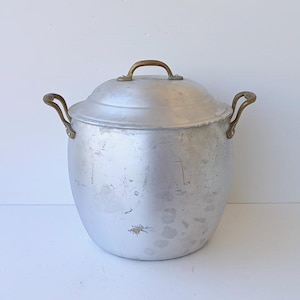 Op de afbeelding: Een grote, ronde soeppan van zilverkleurig metaal met een bijpassend deksel. De pan heeft twee gebogen, messingkleurige handgrepen aan de zijkanten en een enkele boogvormige handgreep op het deksel. Het oppervlak vertoont lichte gebruikssporen.