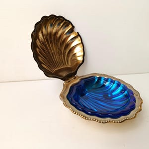 Puede incluir: Una caja de baratijas decorativa en forma de concha. La concha superior es dorada, y la concha inferior es azul vibrante con un patrón ondulado. La caja tiene un borde plateado y está abierta, revelando el interior azul.