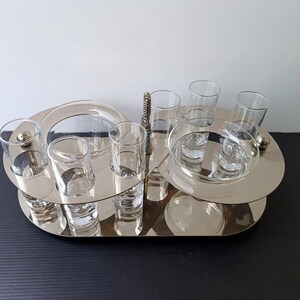 May include: A silver-toned serving tray with multiple levels, holding several clear shot glasses and small bowls. The tray has a unique, layered design with a handle and decorative accents. The glasses and bowls are empty, ready for use.