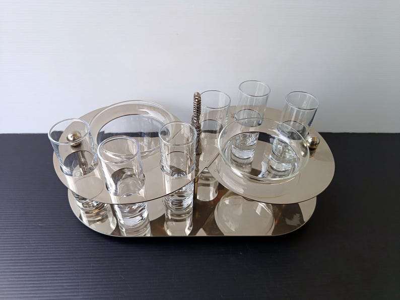 May include: A silver-toned serving tray with multiple levels, holding several clear shot glasses and small bowls. The tray has a unique, layered design with a handle and decorative accents. The glasses and bowls are empty, ready for use.