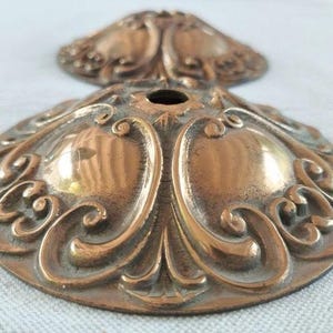 May include: Two circular bronze-toned metal ceiling canopy plates featuring a raised, symmetrical scrollwork pattern and a central circular opening. The metallic surface displays a polished finish with darkened recessed areas highlighting the relief.