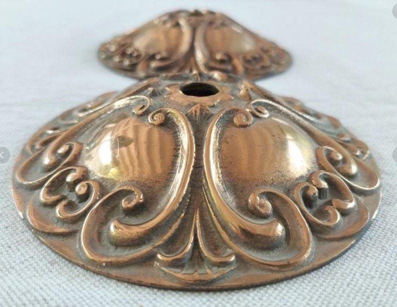 May include: Two circular bronze-toned metal ceiling canopy plates featuring a raised, symmetrical scrollwork pattern and a central circular opening. The metallic surface displays a polished finish with darkened recessed areas highlighting the relief.