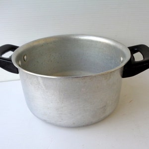 Puede incluir: Una olla de aluminio plateada con asas de plástico negro. La olla está vacía y tiene una superficie lisa y brillante.