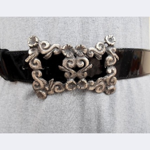 Op de afbeelding: Een zwarte lakleren riem met een grote, rechthoekige zilverkleurige metalen gesp. De gesp heeft een gedetailleerd reliëfontwerp met krullende ornamenten en bloemmotieven, wat zorgt voor een gestructureerde, antiek ogende uitstraling.
