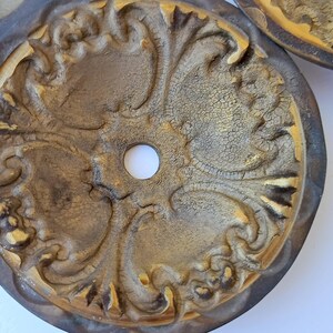 May include: A circular medallion featuring a central hole and an intricate, raised relief pattern of scrolling acanthus leaves. The surface has a distressed, crackled gold finish with dark brown shading in the recessed areas of the design.