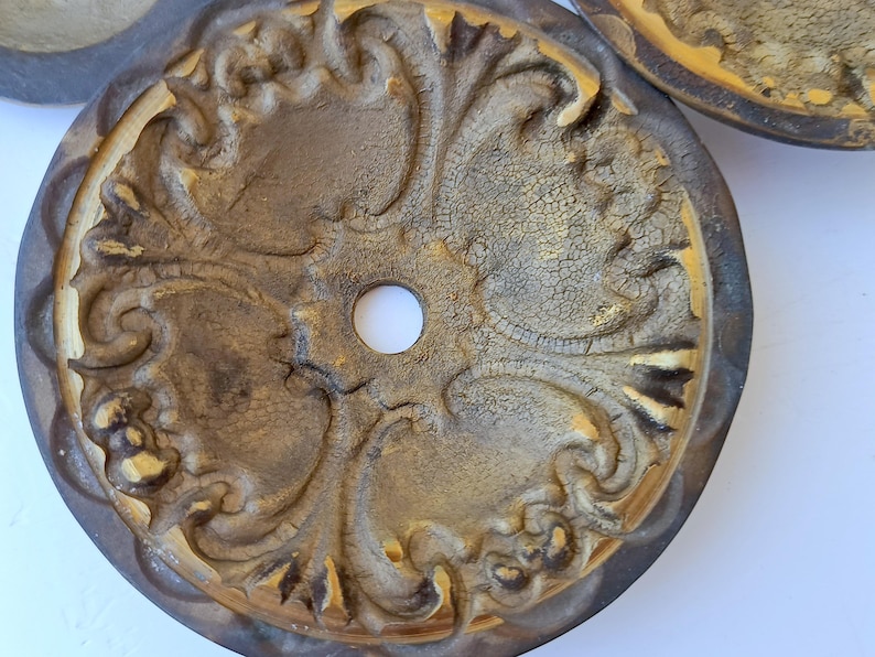 May include: A circular medallion featuring a central hole and an intricate, raised relief pattern of scrolling acanthus leaves. The surface has a distressed, crackled gold finish with dark brown shading in the recessed areas of the design.