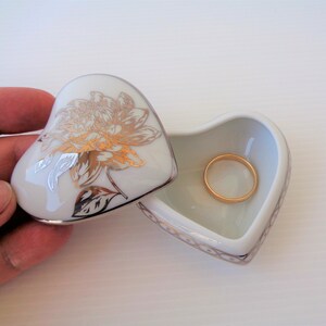 以下が含まれることがあります： ハート型の白いセラミック製の小物入れ。蓋には金色の花の模様が描かれています。箱は開いており、中には金の指輪が入っています。箱の縁は金色の模様で縁取られています。