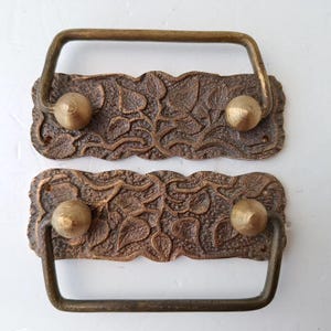 May include: Two rectangular brass drawer pulls featuring a textured, embossed leaf pattern on the backplate. Each pull has a hinged, U-shaped handle attached to two conical mounting points, with small screw holes visible on the edges of the plates.