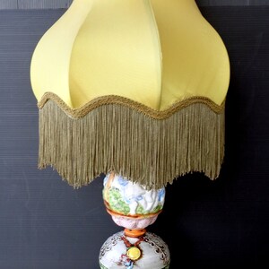 Op de afbeelding: Een vintage tafellamp met een gele lampenkap met een franje rand. De basis is gemaakt van keramiek en heeft een kleurrijk bloemenontwerp met een gouden accent. De lamp staat op een houten basis.