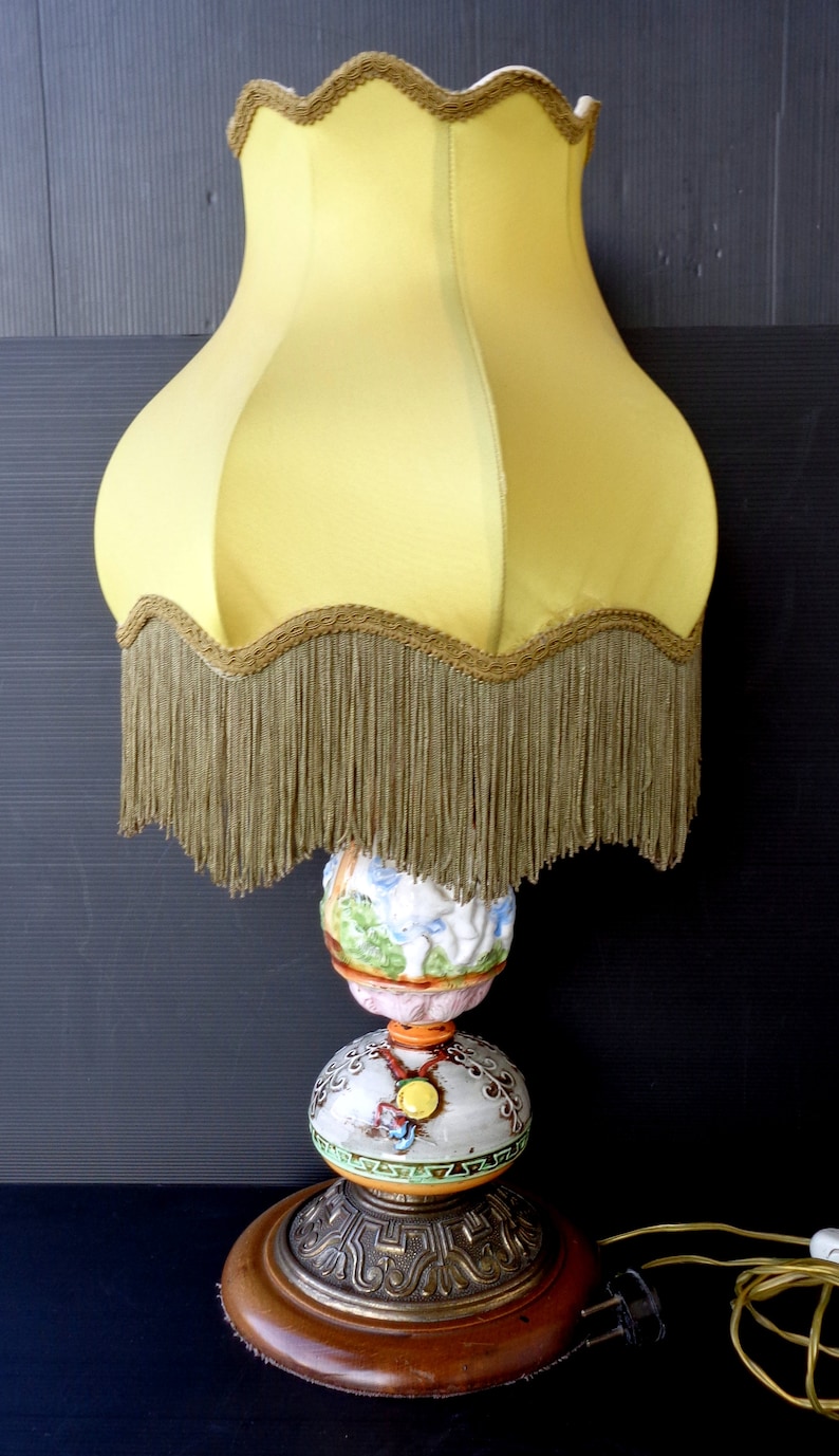 Op de afbeelding: Een vintage tafellamp met een gele lampenkap met een franje rand. De basis is gemaakt van keramiek en heeft een kleurrijk bloemenontwerp met een gouden accent. De lamp staat op een houten basis.