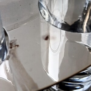 May include: Close-up of clear glass objects, possibly drinking glasses or decorative items, reflecting light. The image has a shallow depth of field, with the focus on the glass surfaces and their reflections. The background is a light, neutral color.
