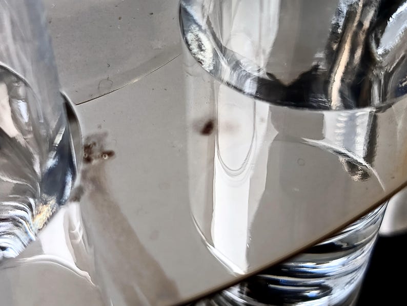 May include: Close-up of clear glass objects, possibly drinking glasses or decorative items, reflecting light. The image has a shallow depth of field, with the focus on the glass surfaces and their reflections. The background is a light, neutral color.
