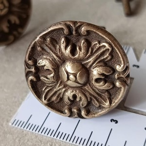 May include: Antique bronze cabinet knob with an ornate floral design. The round knob features a raised central flower surrounded by swirling leaves and decorative flourishes. A ruler is next to the knob, showing measurements in centimetres.
