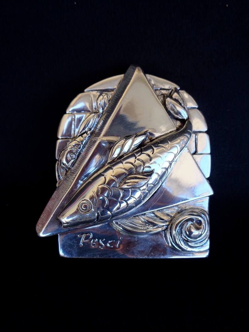May include: Silver-toned metal sculpture of the Pisces zodiac sign. The sculpture features two fish swimming in a triangular frame with the word "Pisces" inscribed below.