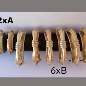 May include: Eight gold-toned metal clasps, arranged in two rows. The top row has two smaller clasps, and the bottom row has six larger clasps. Each clasp features a decorative, ribbed center and a stylized, pointed end. The clasps are displayed on a dark green surface.