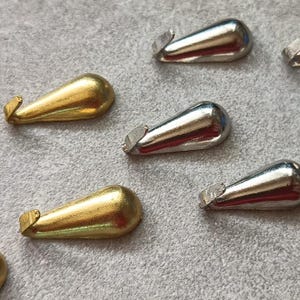 Könnte beinhalten: Nahaufnahme von mehreren tropfenförmigen Metallverschlüssen. Einige sind goldfarben, andere silberfarben. Jeder Verschluss hat einen kleinen Haken an einem Ende, der zum Sichern von Schmuck oder anderen kleinen Gegenständen dient. Die Verschlüsse sind auf einer grauen Oberfläche angeordnet.