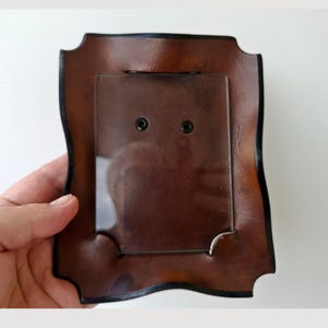 Puede incluir: Un marco de fotos de cuero marrón con un borde negro y una inserción de plástico transparente. El marco tiene una forma decorativa y curvada. La parte posterior del marco es visible, mostrando la inserción y los herrajes de montaje.