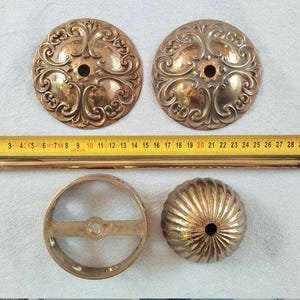 May include: Two circular brass plates with ornate scrollwork, a long thin brass rod, a circular brass ring frame, and a fluted brass dome cap. A yellow measuring tape is positioned horizontally across the items, showing a length of 32 cm.
