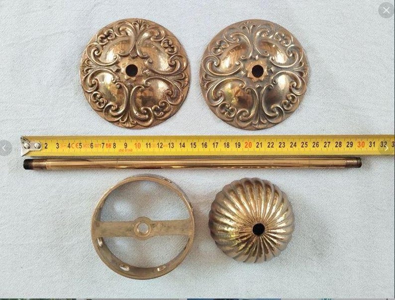 May include: Two circular brass plates with ornate scrollwork, a long thin brass rod, a circular brass ring frame, and a fluted brass dome cap. A yellow measuring tape is positioned horizontally across the items, showing a length of 32 cm.