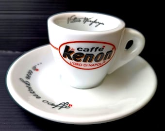 May include: White espresso cup and saucer set. The cup features the text "caffe kenon" in black and red, with "L'ORO DI NAPOLI" below. The saucer has the phrase "...e un gran caffe" in black. The cup has a handle.