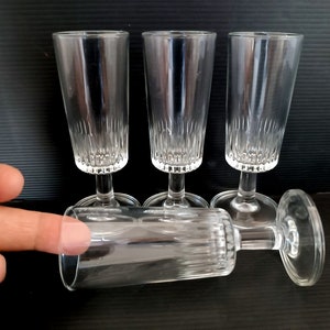 May include: Four clear, stemmed shot glasses with vertical etched lines. Three glasses stand upright, while one lies on its side. The glasses are on a dark surface.