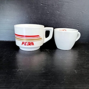 May include: Two white ceramic mugs. The larger mug has a handle and the word "PERA" in red block letters. Above the word are three horizontal stripes in red and gold. The smaller mug is plain white.